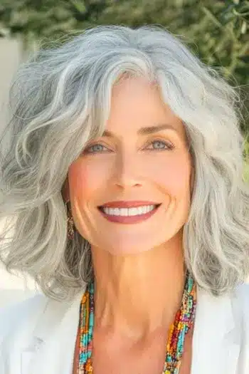 Shoulder-Length Textured Waves with Side-Swept Bangs Haircut on a smiling woman with gray hair.
